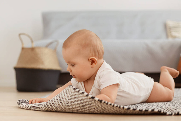 Profile portrait of little tiny infant girl wearing white bodysuit lying on carpet on floor, learning to to crawl at home in light cozy living room, baby playing alone while lying on her belly. - Foto, Bild