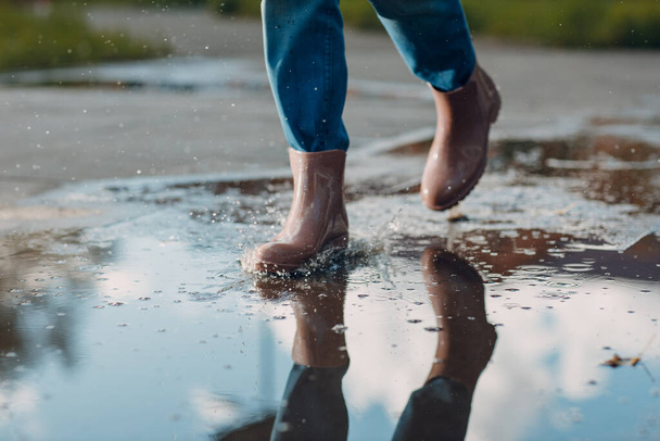 Woman wearing rain rubber boots walking running and jumping into puddle with water splash and drops in autumn rain - Photo, Image