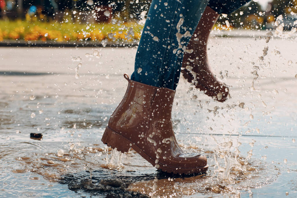 Donna che indossa stivali di gomma antipioggia che cammina correndo e saltando in pozzanghera con spruzzi d'acqua e gocce in autunno pioggia - Foto, immagini