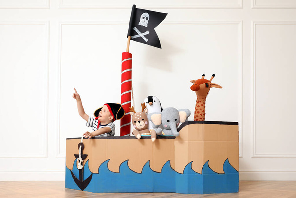 Cute little boy playing with toys in pirate cardboard ship near white wall indoors - Photo, Image