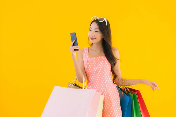 Portrait beautiful young asian woman with colorful shopping bag with smart mobile phone and credit card on yellow isolated background - Fotó, kép