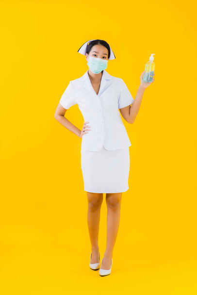 Портрет красива молода азіатка тайська медсестра носить маску для захисту кованої19 або коронавірусу і показує алкогольний гель на жовтому ізольованому фоні
 - Фото, зображення