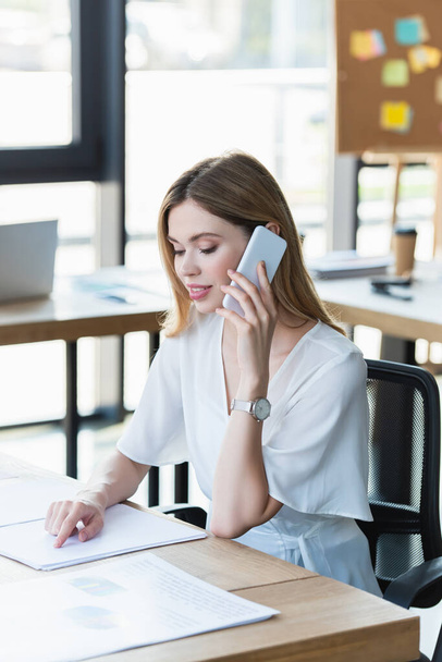 Positive businesswoman talking on smartphone and pointing at document on office table  - Foto, Imagem