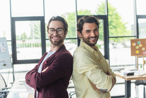 Happy businessmen with crossed arms standing back to back in office  - Fotoğraf, Görsel