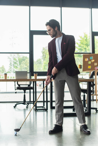 Side view of young businessman in eyeglasses playing golf in office  - 写真・画像
