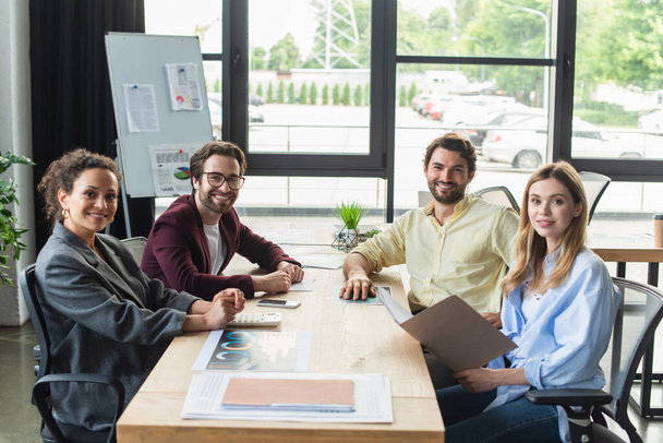 Gente de negocios multicultural positiva mirando la cámara cerca de papeles y calculadora en la oficina  - Foto, Imagen