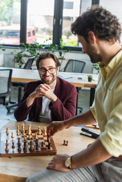 Uomo d'affari sorridente in occhiali guardando collega offuscata giocare a scacchi in ufficio  - Foto, immagini