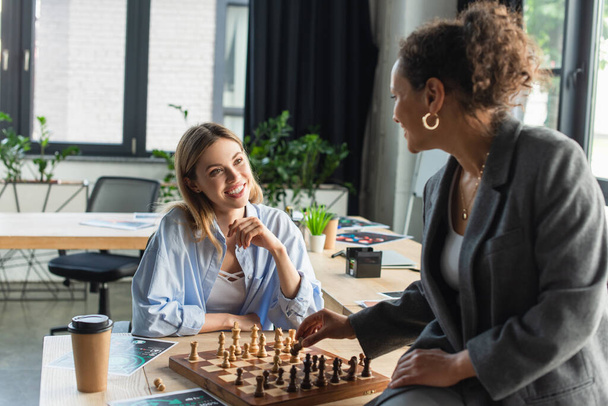 Smiling businesswoman looking at african american colleague playing chess near papers and coffee in office  - Valokuva, kuva