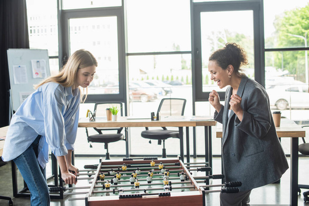 Excited african american businesswoman showing yes gesture while playing table soccer with colleague in office  - Фото, изображение