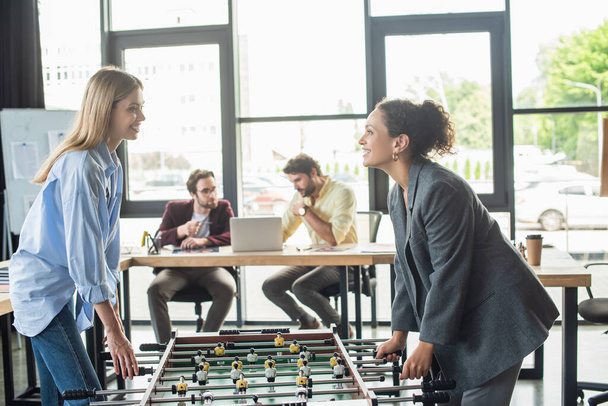 Cheerful businesswomen playing table soccer near blurred businessmen in office  - Фото, изображение