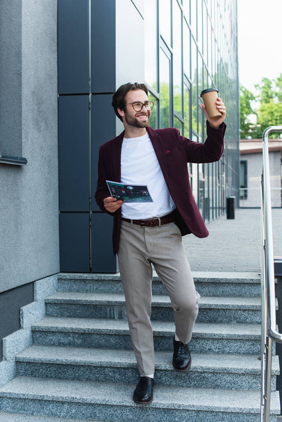 Empresário alegre com copo de papel e documento andando em escadas perto do edifício  - Foto, Imagem