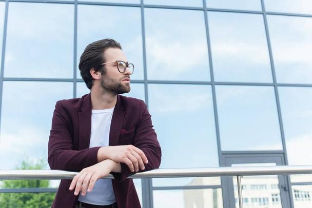 Young businessman in eyeglasses standing near railing and building on urban street  - Foto, Imagem