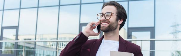 Young businessman with paper folder smiling while talking on smartphone outdoors, banner  - Photo, Image