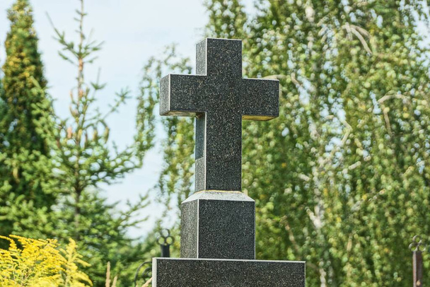 yksi musta kivi risti hautausmaan muistomerkillä sinistä taivasta ja vihreitä puun oksia vasten - Valokuva, kuva