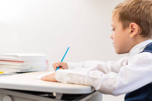 Söpö poika ekaluokkalainen koulupuvussa tekee läksyjä istuessaan pöydän ääressä kynä kädessään kotona. Etäopetus. Valikoiva keskittyminen. Lähemmäs. Muotokuva - Valokuva, kuva