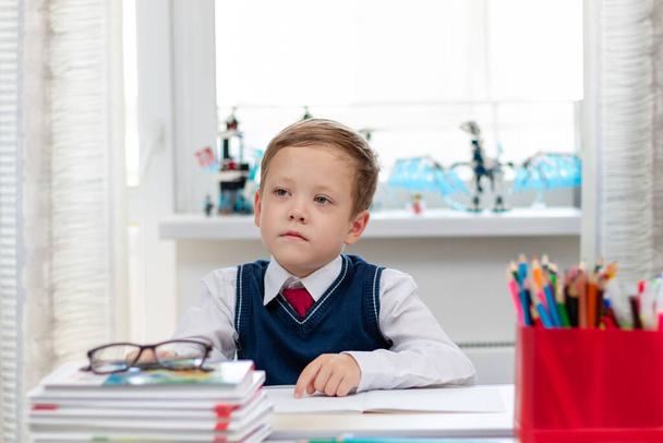 Un garçon mignon de première année dans un uniforme scolaire et une cravate à la maison pendant une pandémie fait ses devoirs alors qu'il est assis à son bureau. Concentration sélective. Gros plan. Portrait - Photo, image