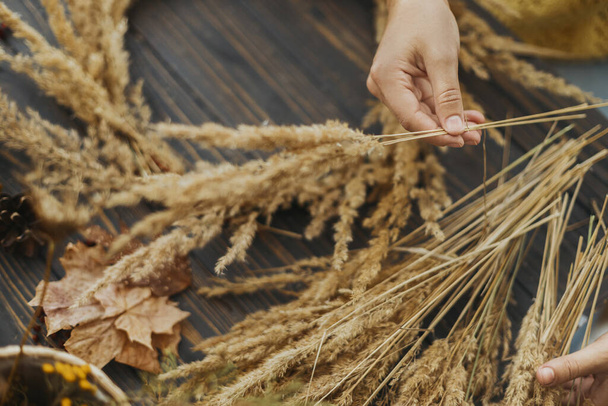 Hands holding dry grass and making stylish wreath with wildflowers, herbs and berries on rustic wooden background. Making rustic autumn boho wreath, holiday workshop - Φωτογραφία, εικόνα