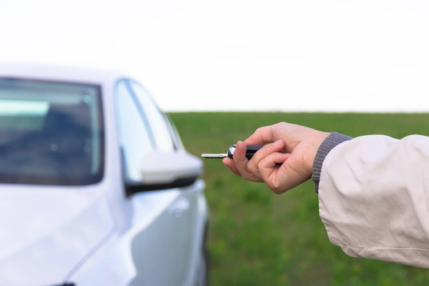 De hand van een vrouw houdt de sleutel van een nieuwe witte auto vast en opent deze op een zonnige zomerdag tegen een achtergrond van groen gras. Selectieve focus. Close-up - Foto, afbeelding