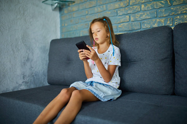 Child using cellphone on the sofa in modern and bright apartment, Young girl plays on smartphone at home, Problem Of gadget Addiction - Fotó, kép