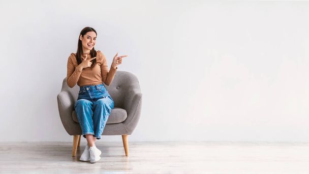 Cool offer. Happy young lady sitting in armchair, pointing aside at free space against white studio wall, banner design - Фото, изображение