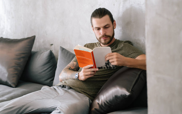 Millennial gutaussehender bärtiger Hipster, der ein Buch hält und liest, während er auf dem Sofa im Café sitzt, entspannt die Zeit im Urlaub. Urlaubsaktivität und Lifestylekonzept. - Foto, Bild