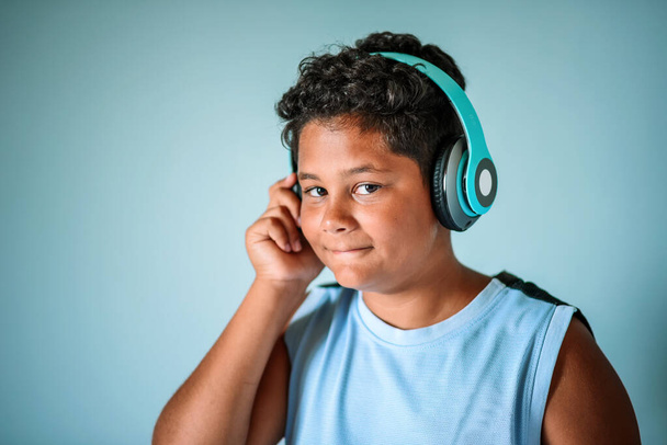 Feliz inteligente encaracolado preteen afro-americano menino desfrutando de música favorita através de fones de ouvido sem fio azuis e olhando para a câmera em estúdio - Foto, Imagem
