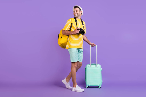 Full body photo of brunet happy positive man walk hold luggage camera casual clothes hat isolated on violet color background - Photo, Image