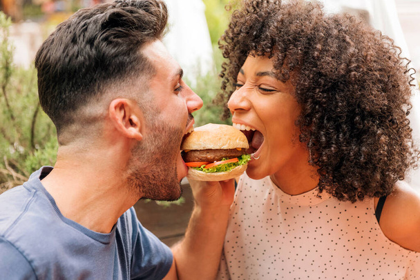 Multiraciální muž a žena jíst chutné hamburger společně během romantického rande v restauraci v létě - Fotografie, Obrázek