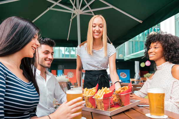 Χαρούμενη χαμογελαστή ξανθιά σερβιτόρα που σερβίρει τηγανητές πατάτες σε ένα τραπέζι παμπ σε μια ομάδα διαφορετικών νεαρών φίλων που απολαμβάνουν μια κρύα μπύρα μαζί - Φωτογραφία, εικόνα