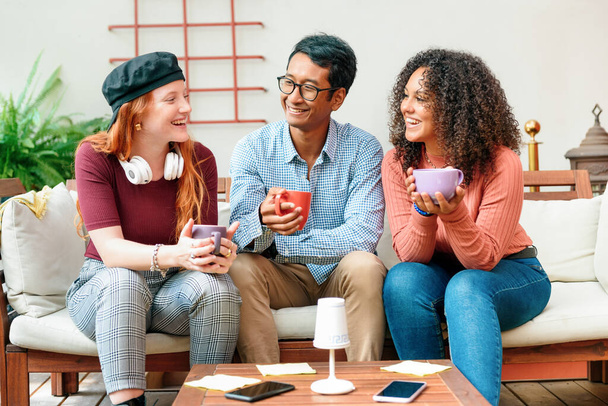 Три различных молодых друга наслаждаются кофе вместе на диване смеясь и улыбаясь, как они проводят некоторое качество время расслабляясь и охлаждения в современной гостиной - Фото, изображение
