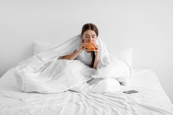 Λυπηρή πεινασμένη νεαρή γυναίκα κάθεται στο λευκό κρεβάτι στο σπίτι και τρώει burger, υποφέρει από κατάθλιψη και ψυχικά προβλήματα - Φωτογραφία, εικόνα