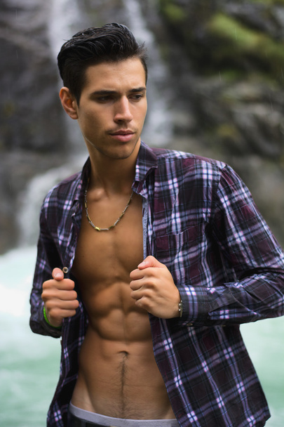 Handsome young man near mountain waterfall with open shirt - Photo, Image