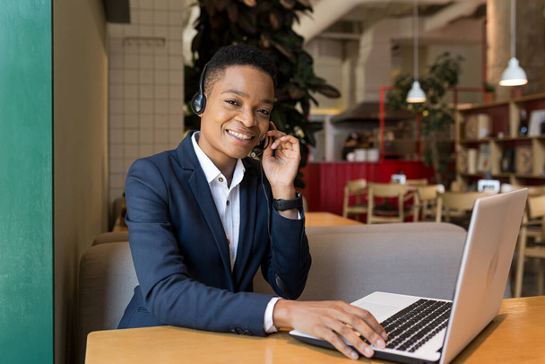 Beautiful african american woman looking at camera and smiling, working in coworking cafe, video communication using headset business woman freelancer - Fotó, kép