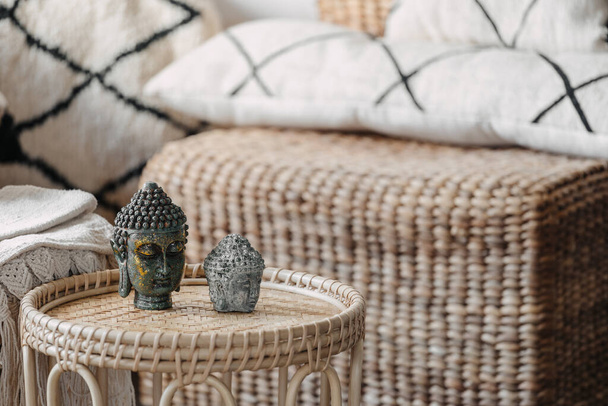 Close up view of Buddha head standing on wicked coffee table. Home decor on bamboo furniture with natural materials in bright living room interior at bohemian style. Calmness and mental health concept - Photo, Image