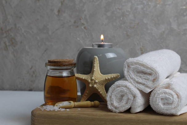 Wellness-Massage Körperpflege zu Hause. Weiße Handtücher Öl duftend für Massage Aromatherapie Kerzen Stern Meer liegen auf einem Holztablett auf einem grauen Hintergrund Seitenansicht. - Foto, Bild
