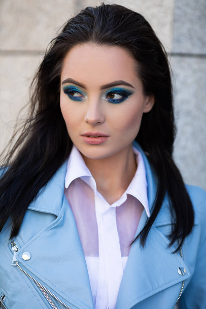 Portrait de jeune femme brune aux cheveux longs et maquillage vêtu d'une veste bleue dans la rue. - Photo, image