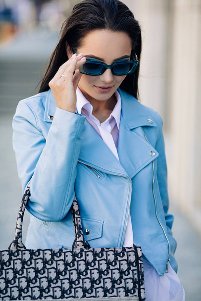 Young brunette woman in sunglasses stands on street dressed in blue jacket with bag on her hand. Portrait. - Photo, Image