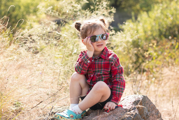 Маленькая улыбающаяся рыжая девочка в солнечных очках и хвостиках на размытом деревенском фоне. Симпатичный красивый ребенок в красной коричневой рубашке сидит на большом камне на размытом фоне - Фото, изображение