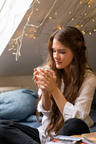 Young woman sitting on bed dreaming and drinking hot water or tea at home - Foto, Imagem