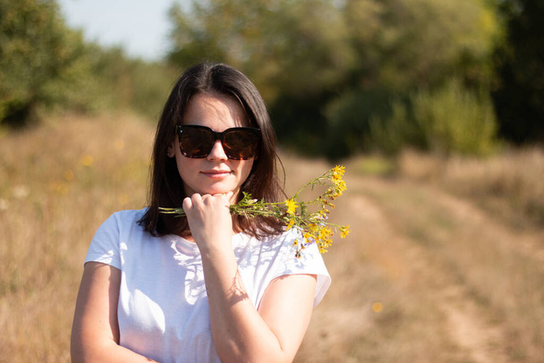Una joven hermosa chica blanca con el pelo largo y oscuro y gafas de sol se encuentra en el fondo de un camino de pueblo - Foto, Imagen