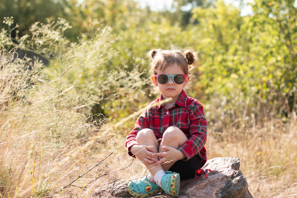 Маленька усміхнена руда дівчинка в сонцезахисних окулярах і хвостах на розмитому сільському тлі. Симпатична красива дитина в червоно-коричневій сорочці сидить на великому камені на розмитому тлі
 - Фото, зображення
