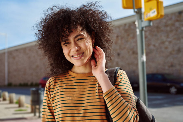 Smiling young black woman touching her hair while standing with a backpack outdoors during the day - 写真・画像