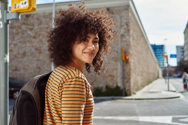 Cute young black woman looking at the camera cheerfully while crossing a street outdoors during the day - Photo, image