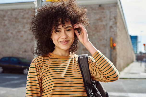 Cute young black woman looking at the camera cheerfully while touching her hair and carrying a backpack outdoors during the day - Foto, immagini