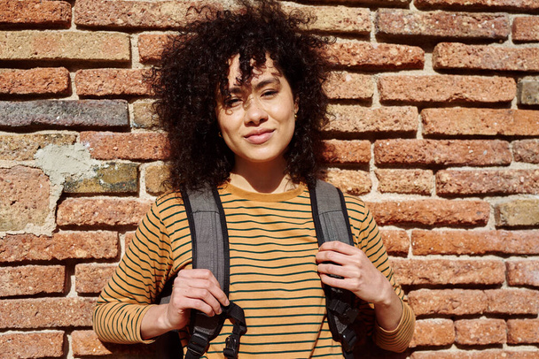 Cute young black woman looking at the camera while standing against a brick wall with a backpack during the day - Zdjęcie, obraz