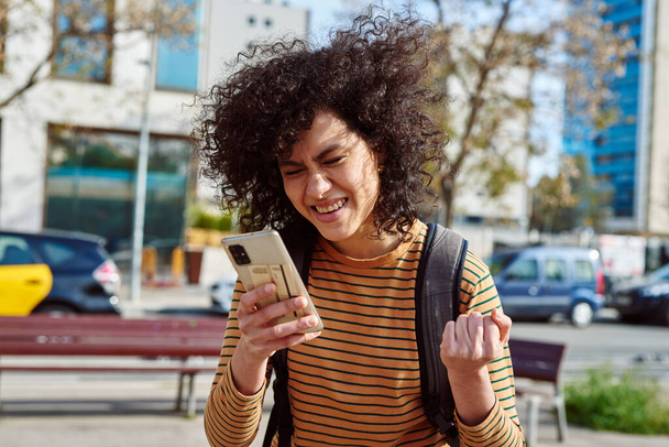 Cheerful young black woman with a backpack celebrating while using a cellphone in the city during the day - Fotografie, Obrázek