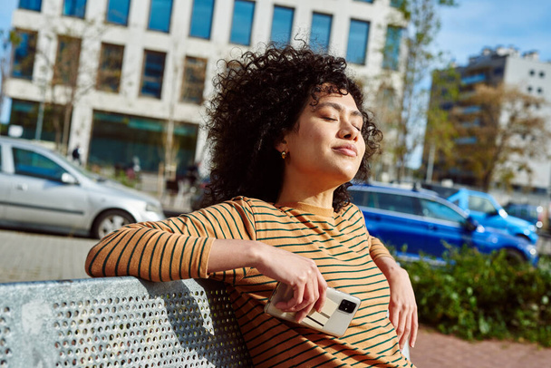 Cute young black woman holding a cellphone while sitting on a bench with her eyes closed outdoors in the city - Foto, afbeelding