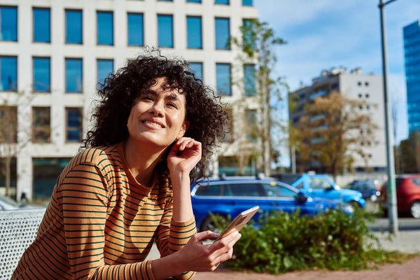 Smiling young black woman looking away cheerfully while holding a cellphone in her hand outdoors in the city - 写真・画像