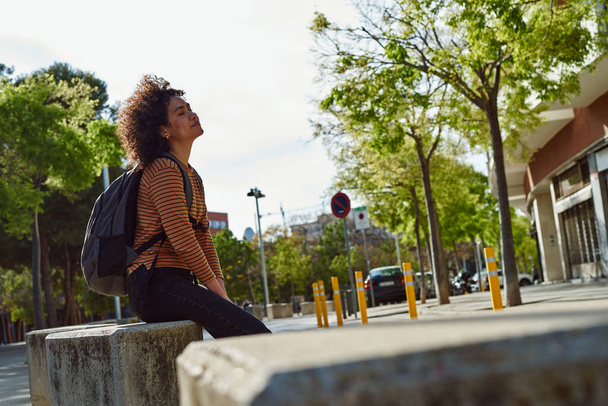 Cute young black woman with a backpack relaxing on a stone slab with her eyes closed outdoors during the day - Photo, Image