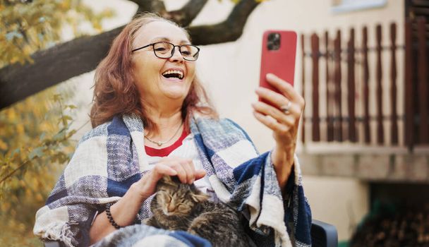 Beautiful senior happy smiling woman chatting using smartphone online with cat in hands in autumn garden of her country house - Fotoğraf, Görsel